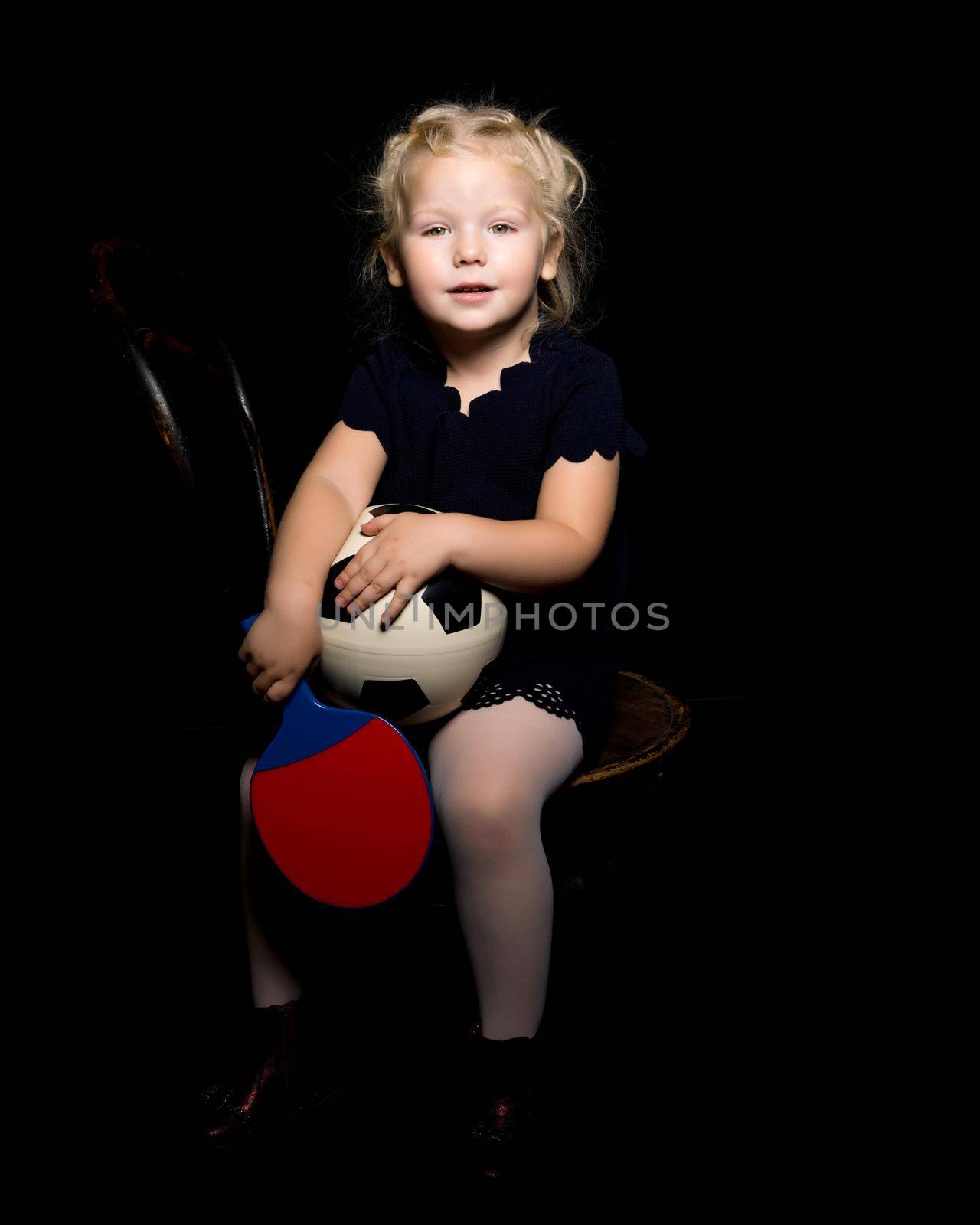 A cute little girl is playing with a soccer ball. Studio photo on a black background. The concept of a happy childhood, sport and fashion.