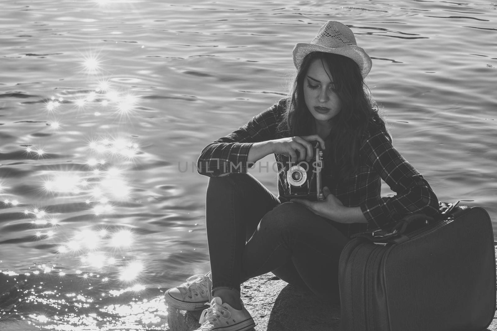 Young woman taking photographs with a vintage camera sitting in a harbour. Girl wearing plaid shirt, blue jeans and sun hat. Black and white photograph.