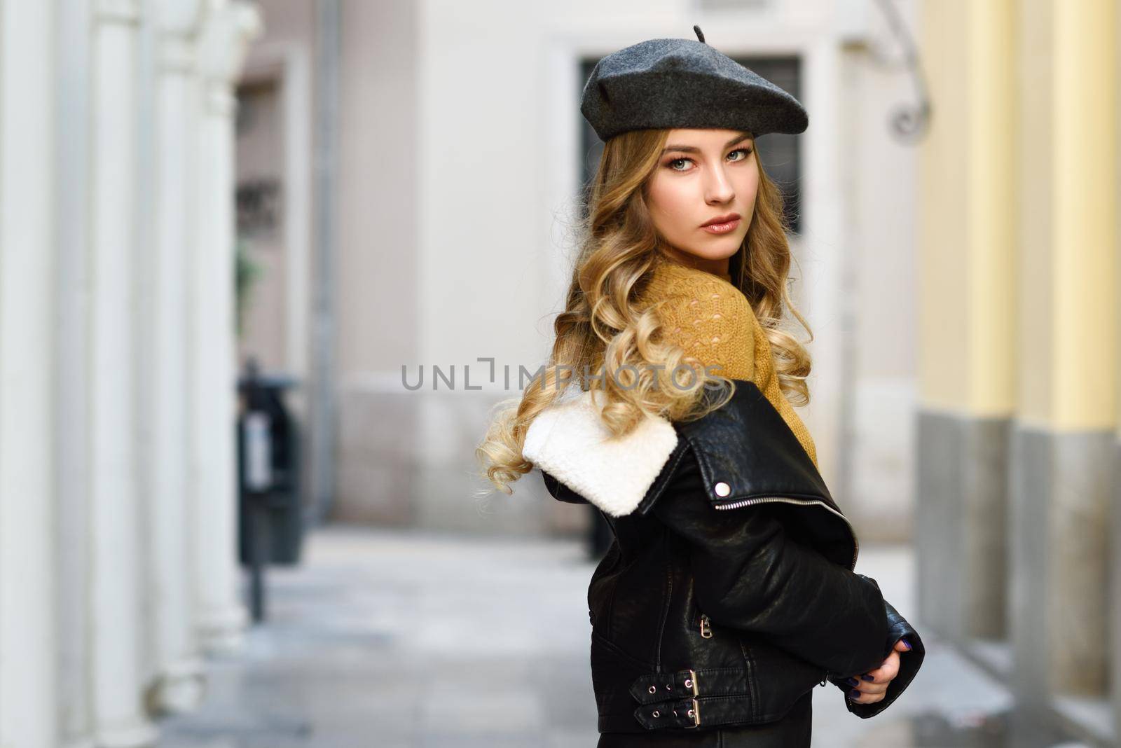 Blonde russian woman in urban background. Beautiful young girl wearing beret, black leather jacket and mini skirt standing in the street. Pretty female with long wavy hair hairstyle and blue eyes.