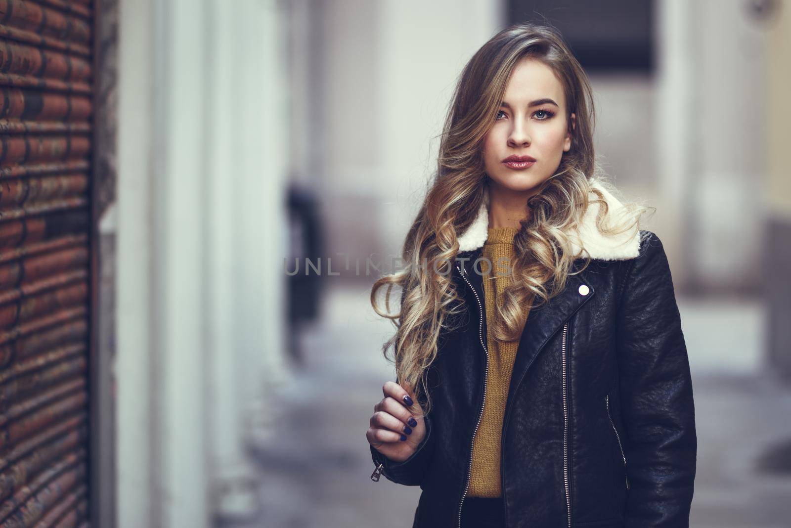 Blonde woman in urban background. Beautiful young girl wearing black leather jacket and mini skirt standing in the street. Pretty russian female with long wavy hair hairstyle and blue eyes.