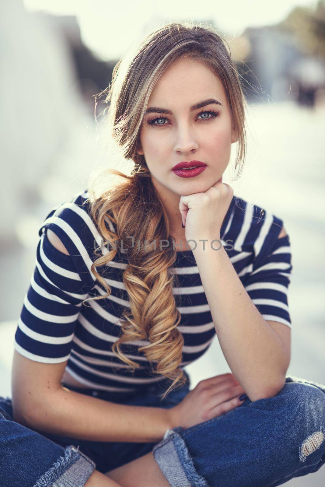 Blonde woman, model of fashion, sitting on a bench in urban background. Beautiful young girl wearing striped t-shirt and blue jeans in the street. Pretty russian female with pigtail.