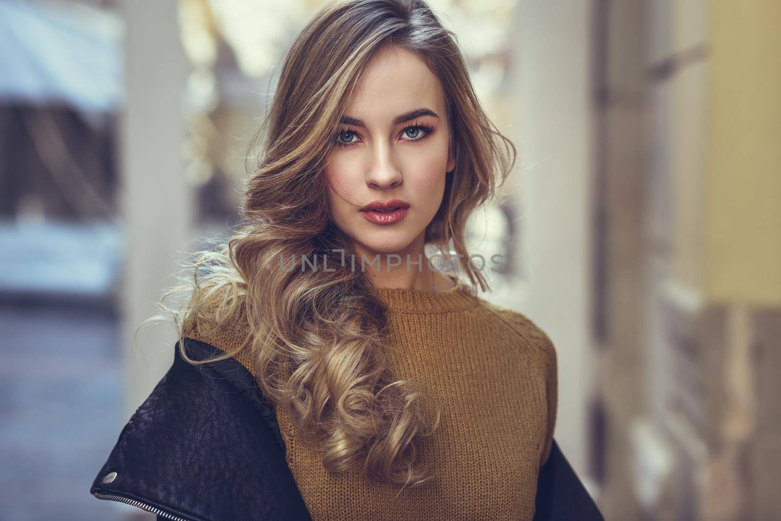 Blonde woman in urban background. Beautiful young girl wearing black leather jacket and mini skirt standing in the street. Pretty russian female with long wavy hair hairstyle and blue eyes.
