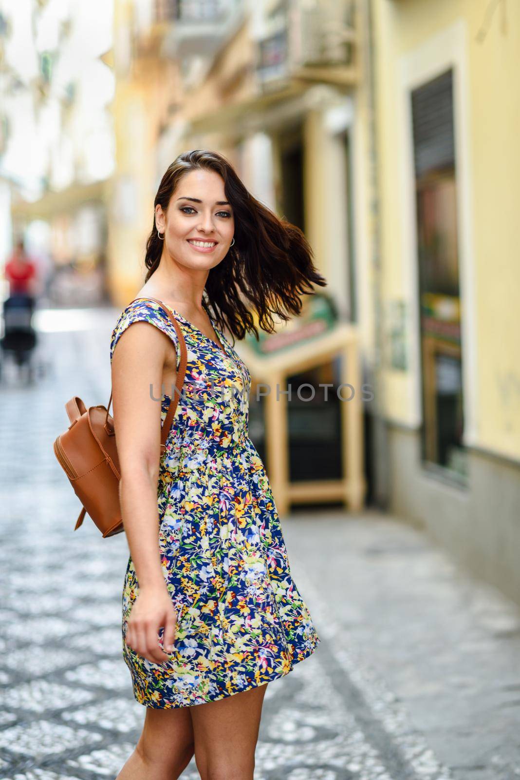 Smiling young woman with blue eyes with brown wavy hair outdoors. Girl wearing flower dress in urban background. Beauty and fashion concept.