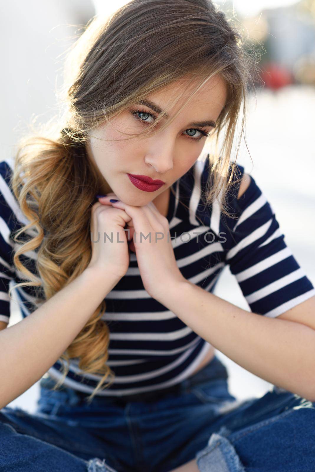 Blonde woman, model of fashion, sitting on a bench in urban background. Beautiful young girl wearing striped t-shirt and blue jeans in the street. Pretty russian female with pigtail.