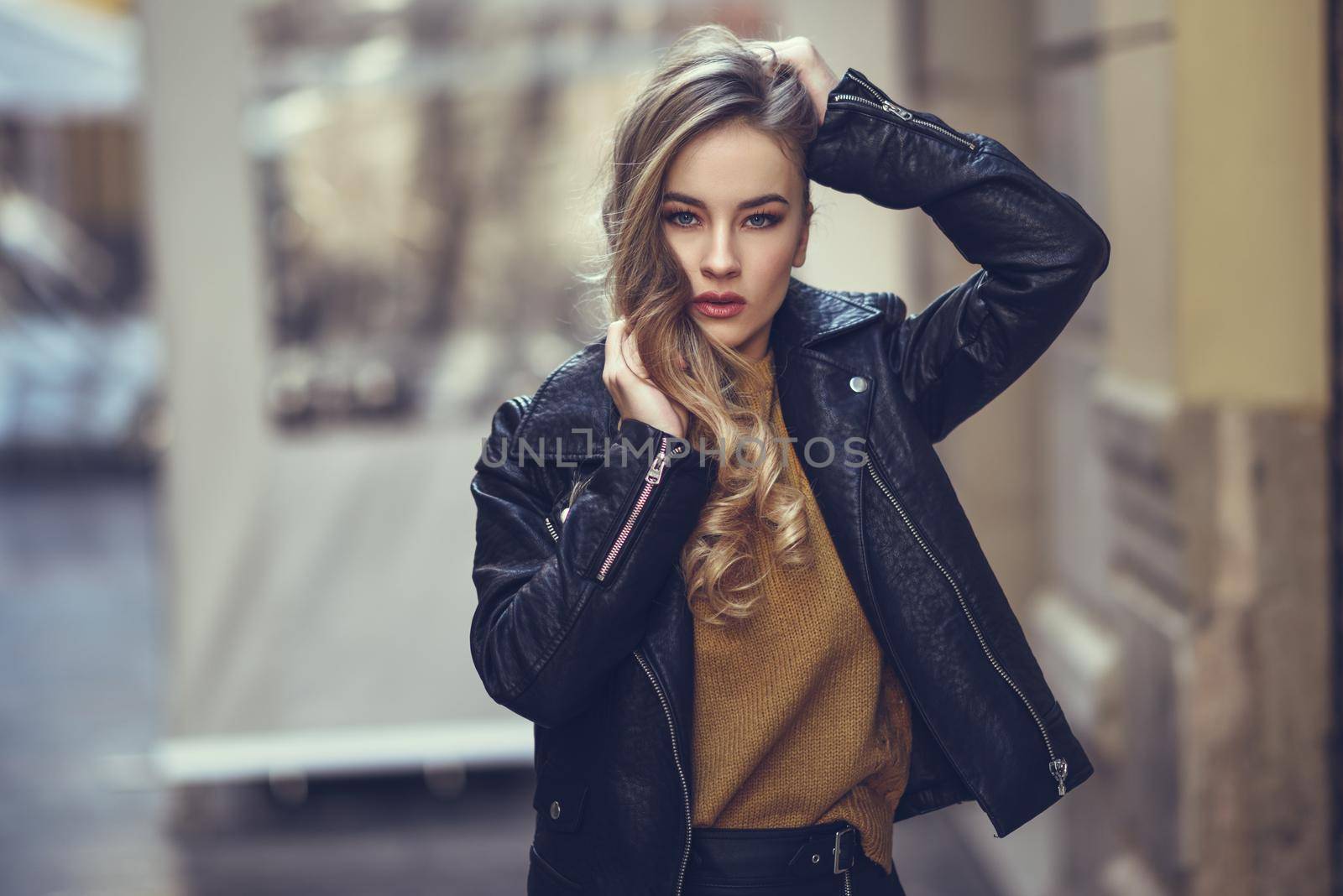 Blonde woman in urban background. Beautiful young girl wearing black leather jacket and mini skirt standing in the street. Pretty russian female with long wavy hair hairstyle and blue eyes.