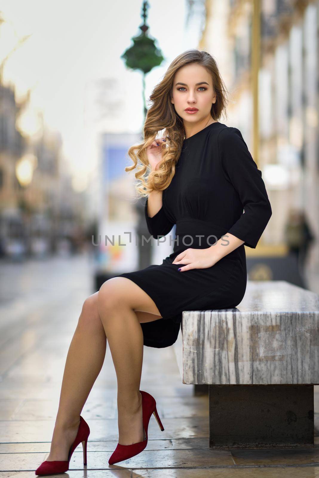 Blonde woman in urban background. Beautiful young girl wearing black elegant dress and red high heels standing in the street. Pretty russian female with long wavy hair hairstyle and blue eyes.