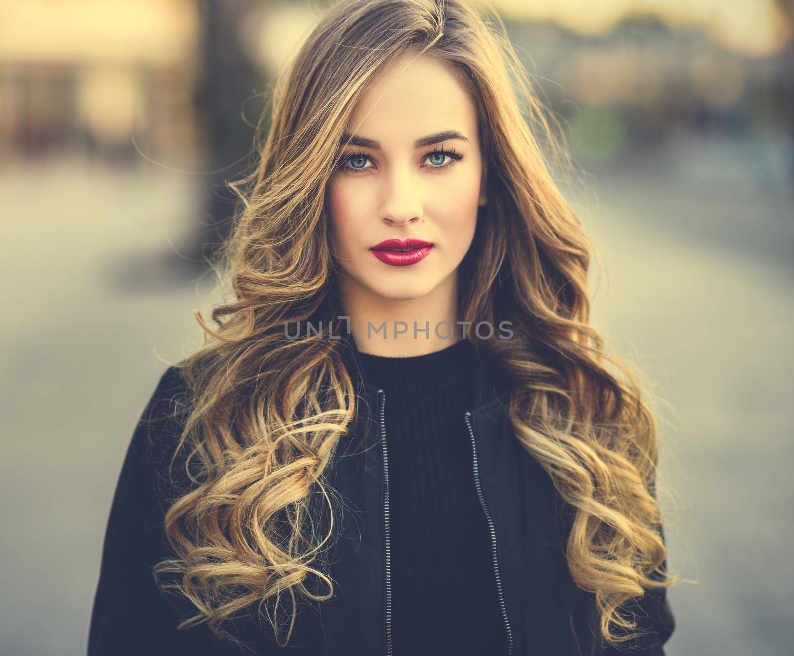 Close-up portrait of young blonde girl with beautiful blue eyes wearing black jacket outdoors. Pretty russian female with long wavy hair hairstyle. Woman in urban background.