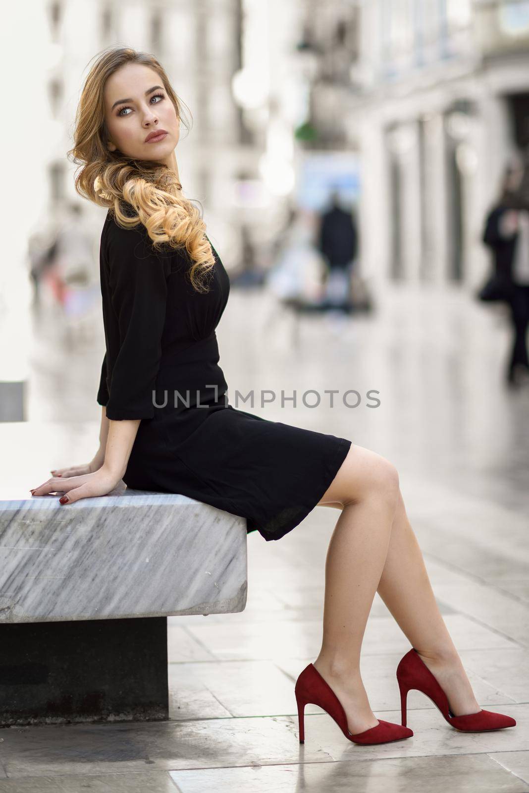 Blonde woman with beautiful legs in urban background. Beautiful young girl wearing black elegant dress and red high heels sitting on a bench in the street. Pretty russian female with long wavy hair hairstyle and blue eyes.