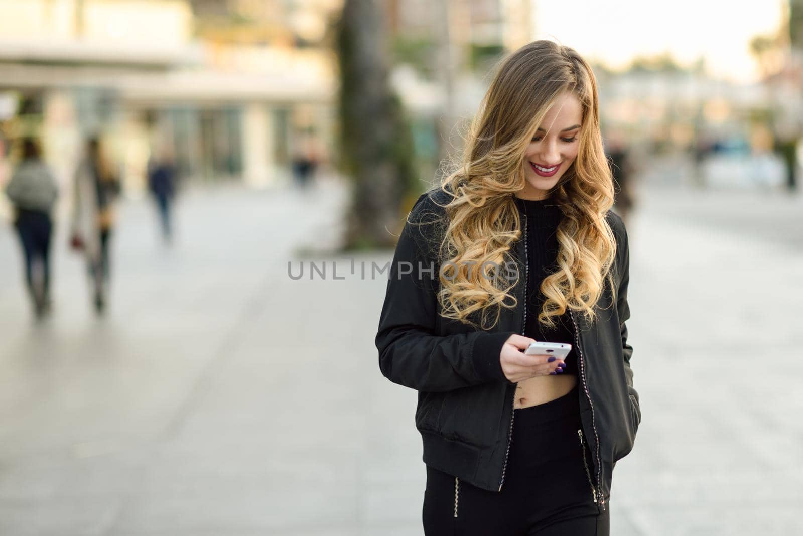 Blonde woman texting with her smart phone in urban background. Beautiful young girl wearing black jacket walking in the street. Pretty russian female with long wavy hair hairstyle.