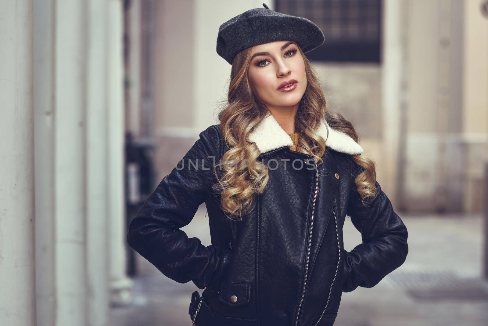 Blonde russian woman in urban background. Beautiful young girl wearing beret, black leather jacket and mini skirt standing in the street. Pretty female with long wavy hair hairstyle and blue eyes.