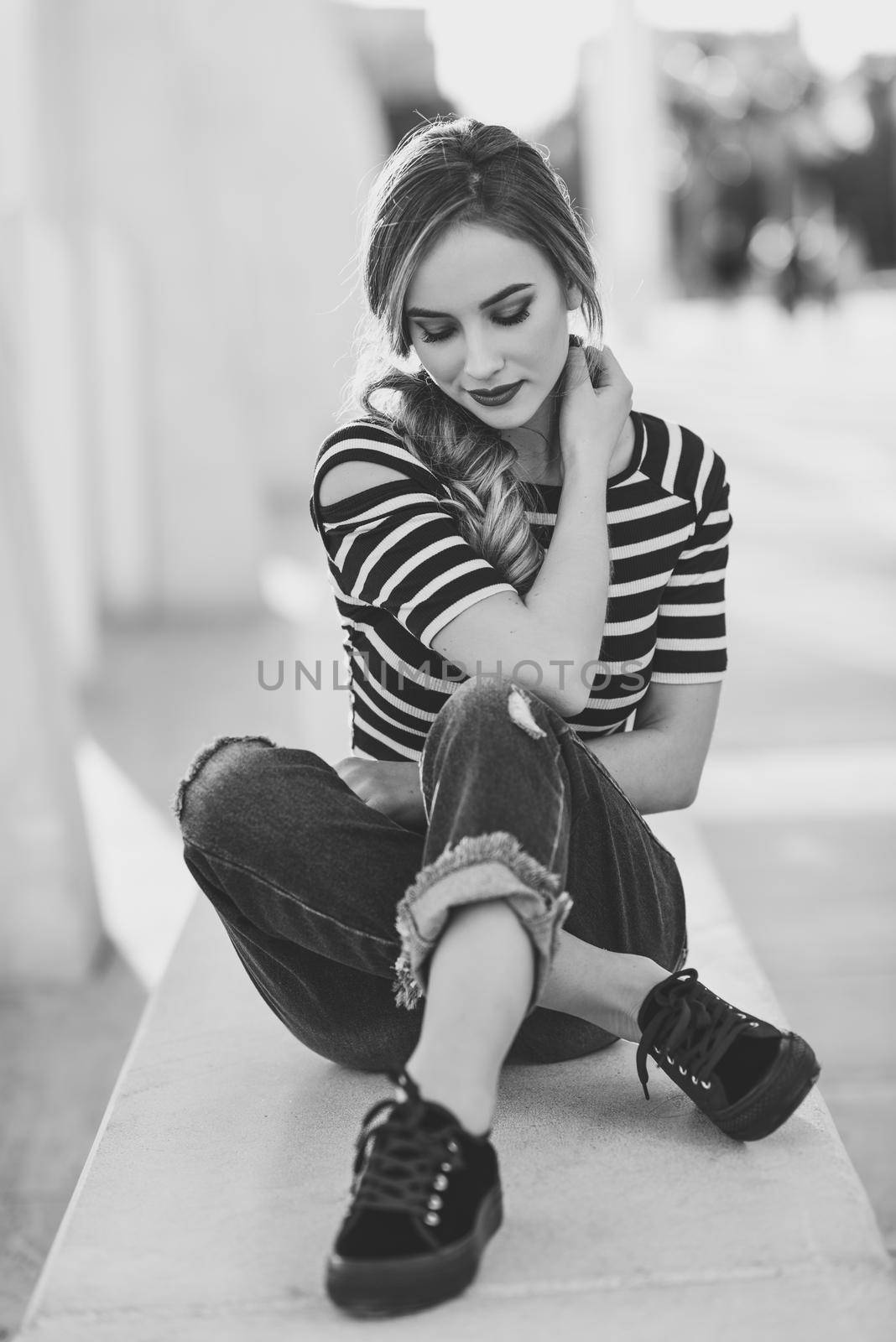 Blonde woman, model of fashion, sitting on a bench in urban background with eyes closed. Thoughtful young girl wearing striped t-shirt and blue jeans in the street. Pretty russian female with pigtail.