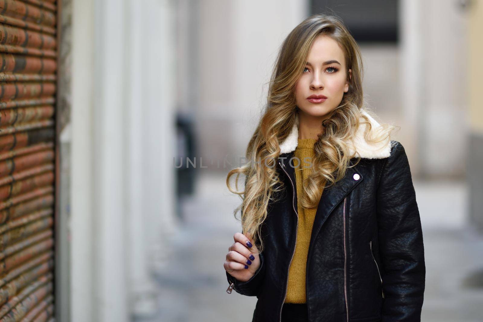 Blonde woman in urban background. Beautiful young girl wearing black leather jacket and mini skirt standing in the street. Pretty russian female with long wavy hair hairstyle and blue eyes.