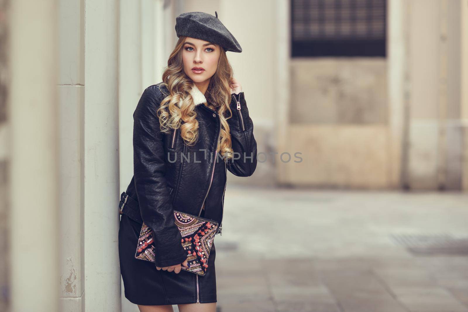 Blonde russian woman in urban background. Beautiful young girl wearing beret, black leather jacket and mini skirt standing in the street. Pretty female with long wavy hair hairstyle and blue eyes.