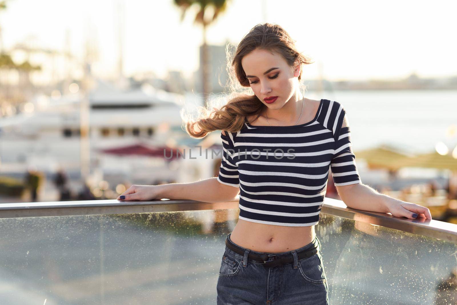 Blonde woman, model of fashion, standing in a harbor with boats and sea in the background. Beautiful young girl wearing striped t-shirt and blue jeans. Pretty russian female with pigtail.