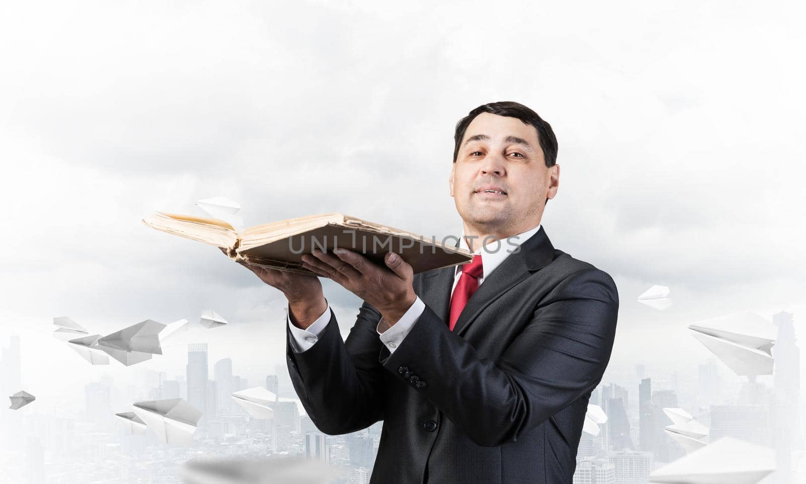 Serious businessman holding open book. Man in business suit standing on cloudy cityscape background with flying paper planes. Lawyer reading legal regulation book. Business accounting and consulting