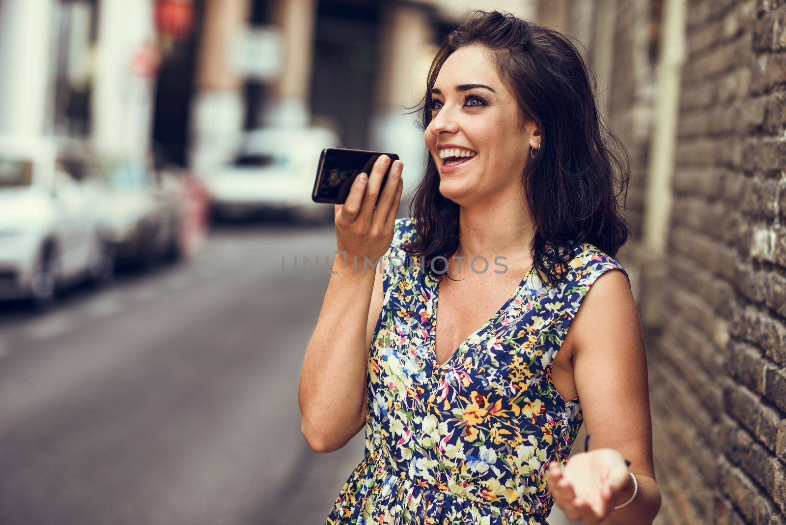 Smiling young woman recording voice note in her smart phone outdoors. Girl wearing flower dress in urban background. Technology concept.