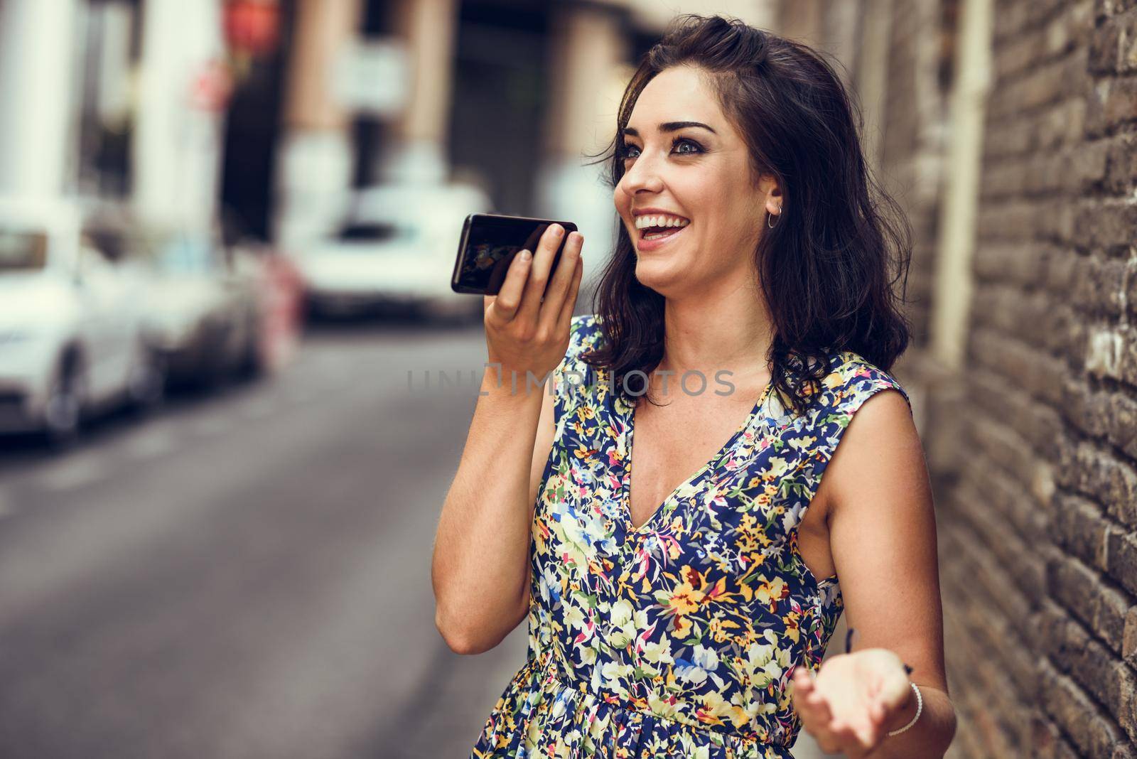 Smiling young woman recording voice note in her smart phone outdoors. Girl wearing flower dress in urban background. Technology concept.