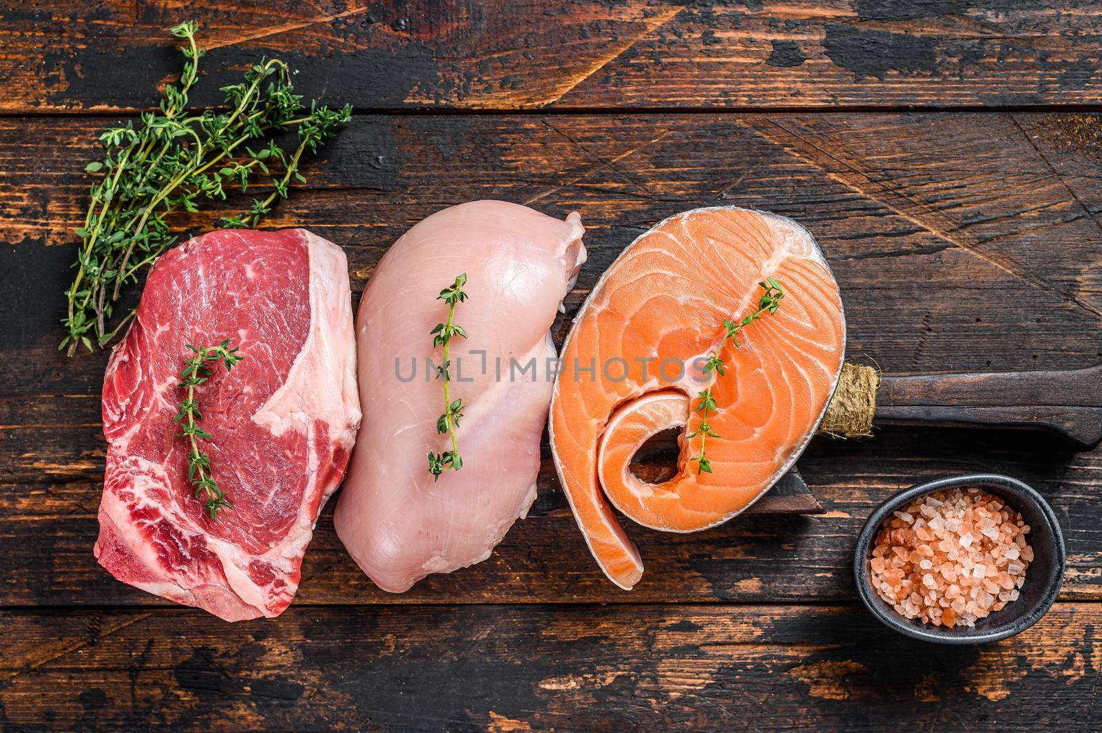 Different types of raw meat steaks Beef striploin, salmon and chicken breast. Dark wooden background. Top view.