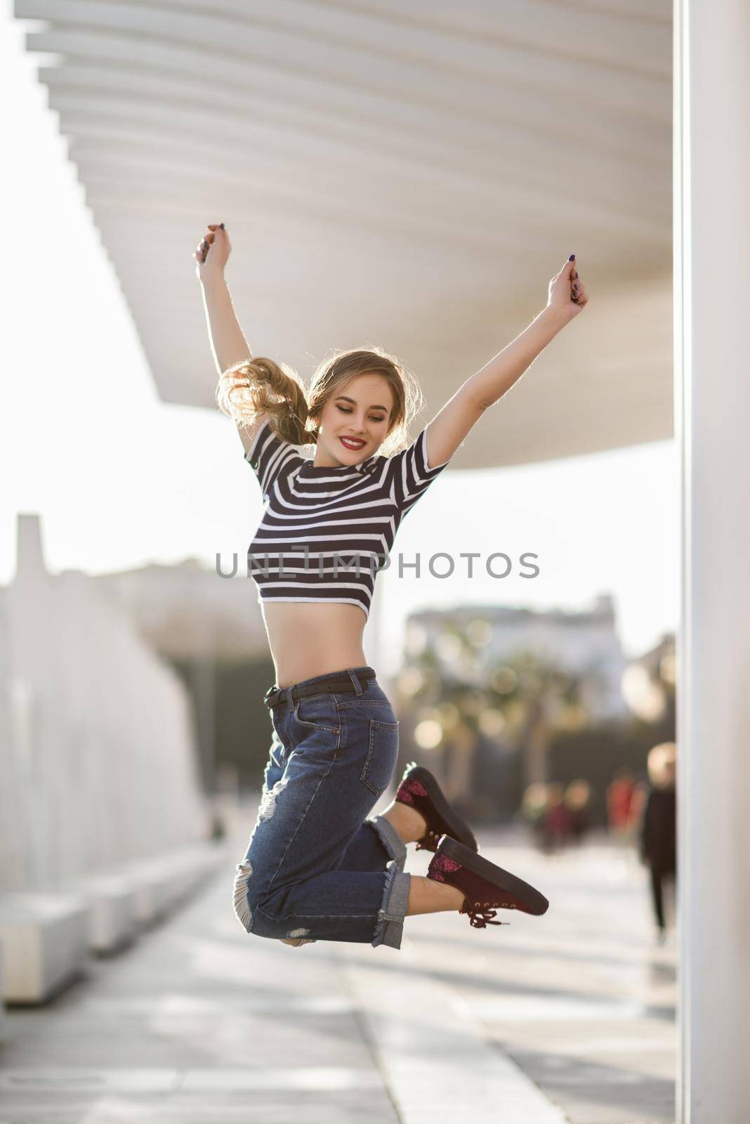 Funny blonde woman jumping in urban background. Beautiful young girl wearing striped t-shirt and blue jeans in the street. Pretty russian female with pigtail.