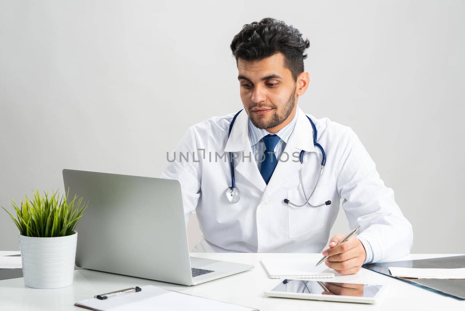Handsome doctor sitting in office. Young practitioner in white coat with stethoscope at desk with laptop. Professional medical consultation and treatment in modern clinic. Medical healthcare concept