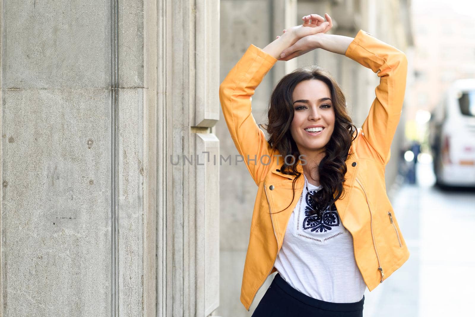 Young brunette woman, model of fashion, wearing orange modern jacket and blue skirt. Pretty caucasian girl with long wavy hairstyle smiling. Female raising her arms in urban background.