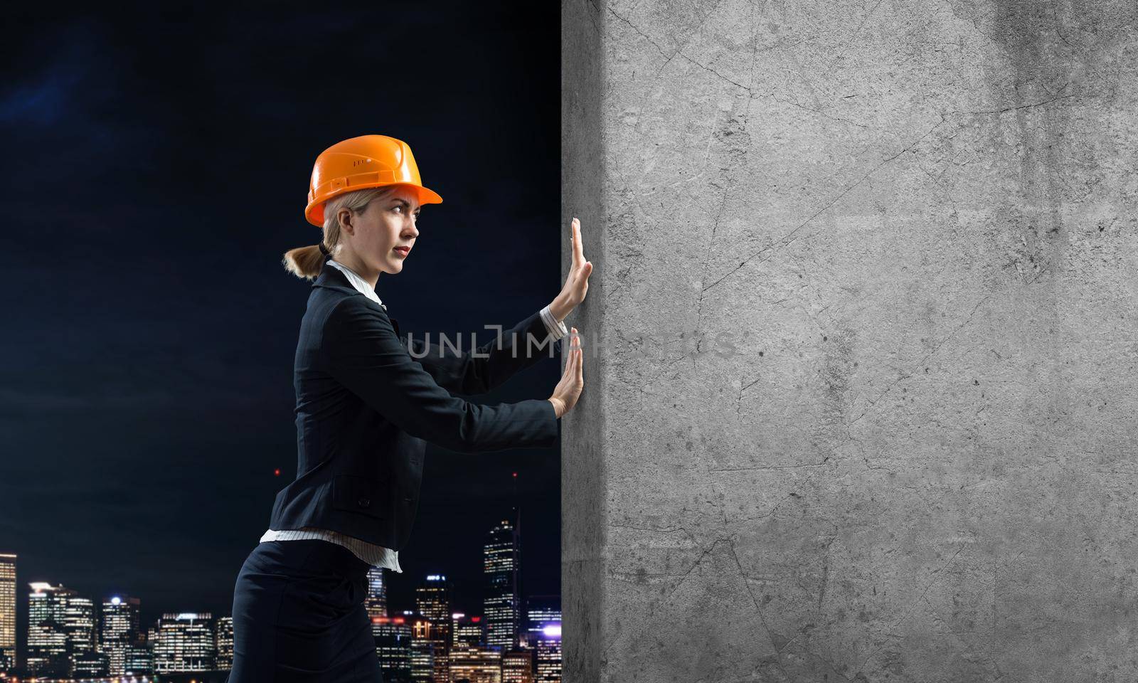 Beautiful woman architect pushing concrete wall. Young specialist in safety helmet on night city background. Overcoming obstacles and solution of problems. Grey concrete wall as banner with copy space