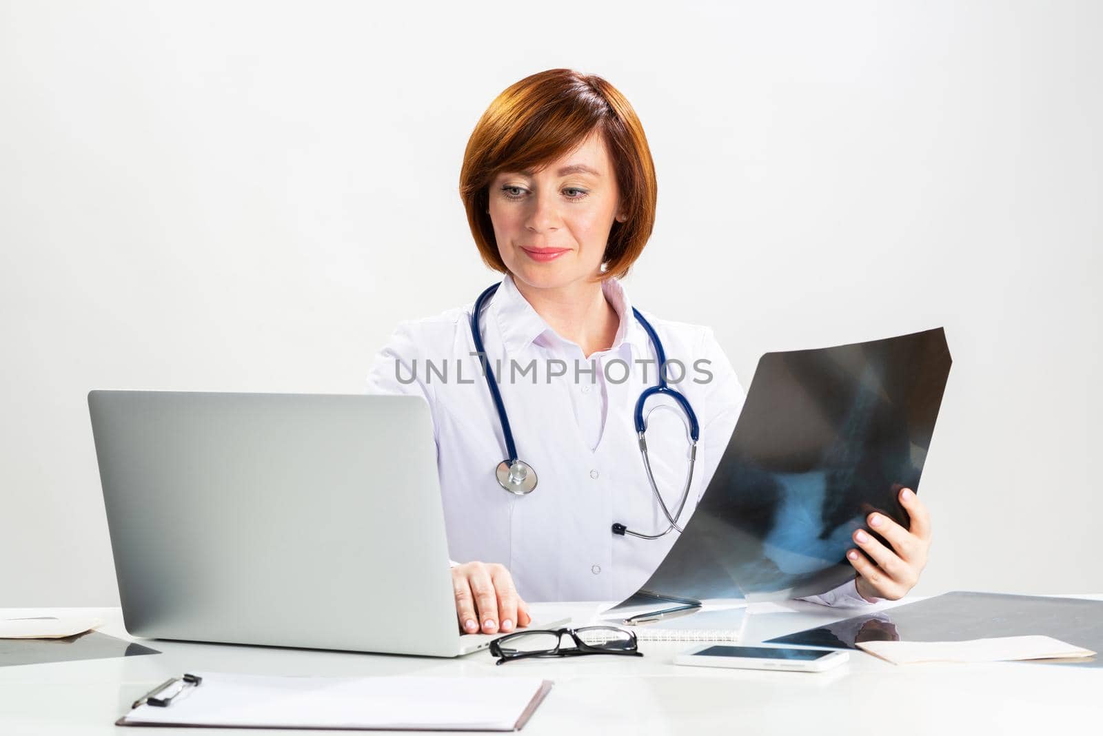 Beautiful doctor looking at x-ray scan in office. Physician in white coat with stethoscope sitting at desk with laptop. Professional medical diagnosis and treatment in clinic. Medical check up service