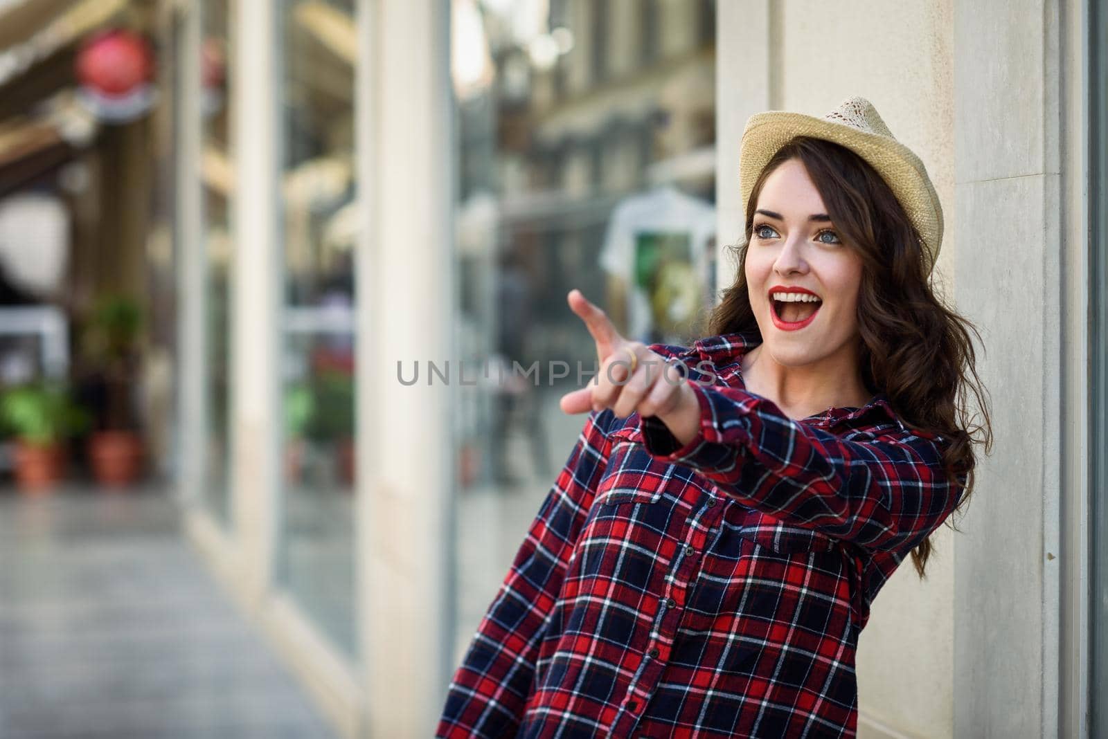 Young woman with beautiful blue eyes wearing plaid shirt and sun hat pointing with her finger. Girl in urban background.