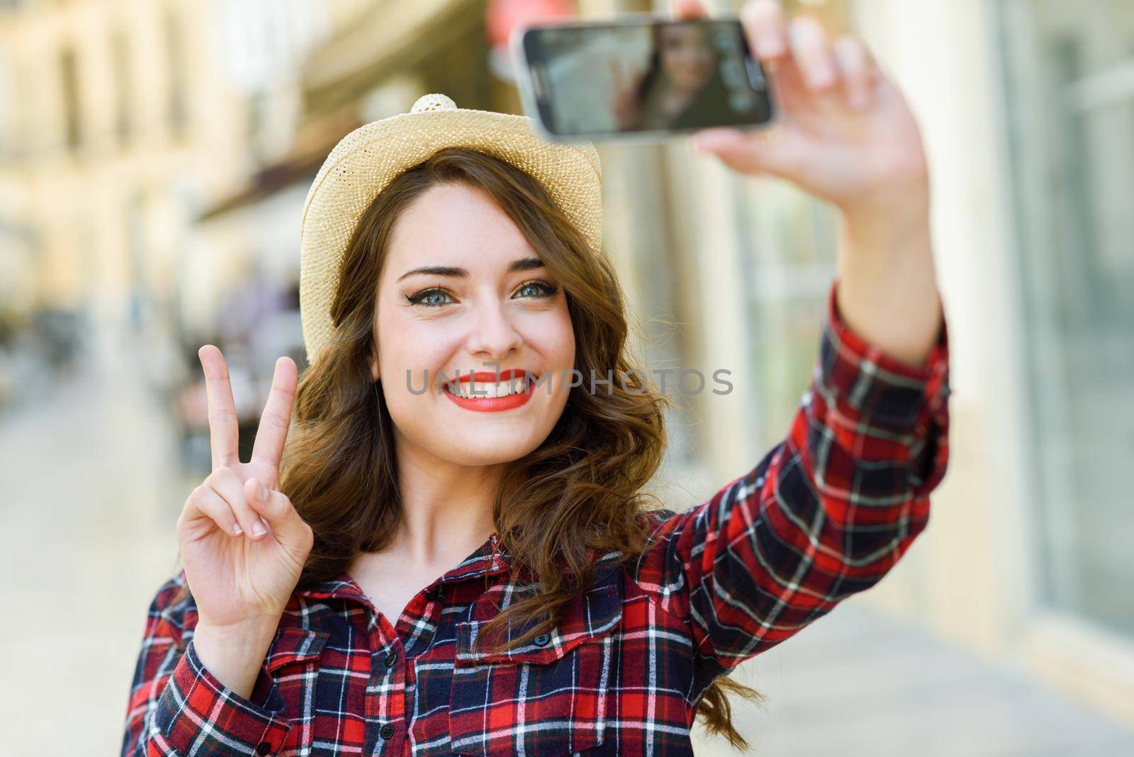 Portrait of a beautiful young woman selfie in the street with a smartphone. Young woman wearing plaid shirt and sun hat with red lips in urban background.