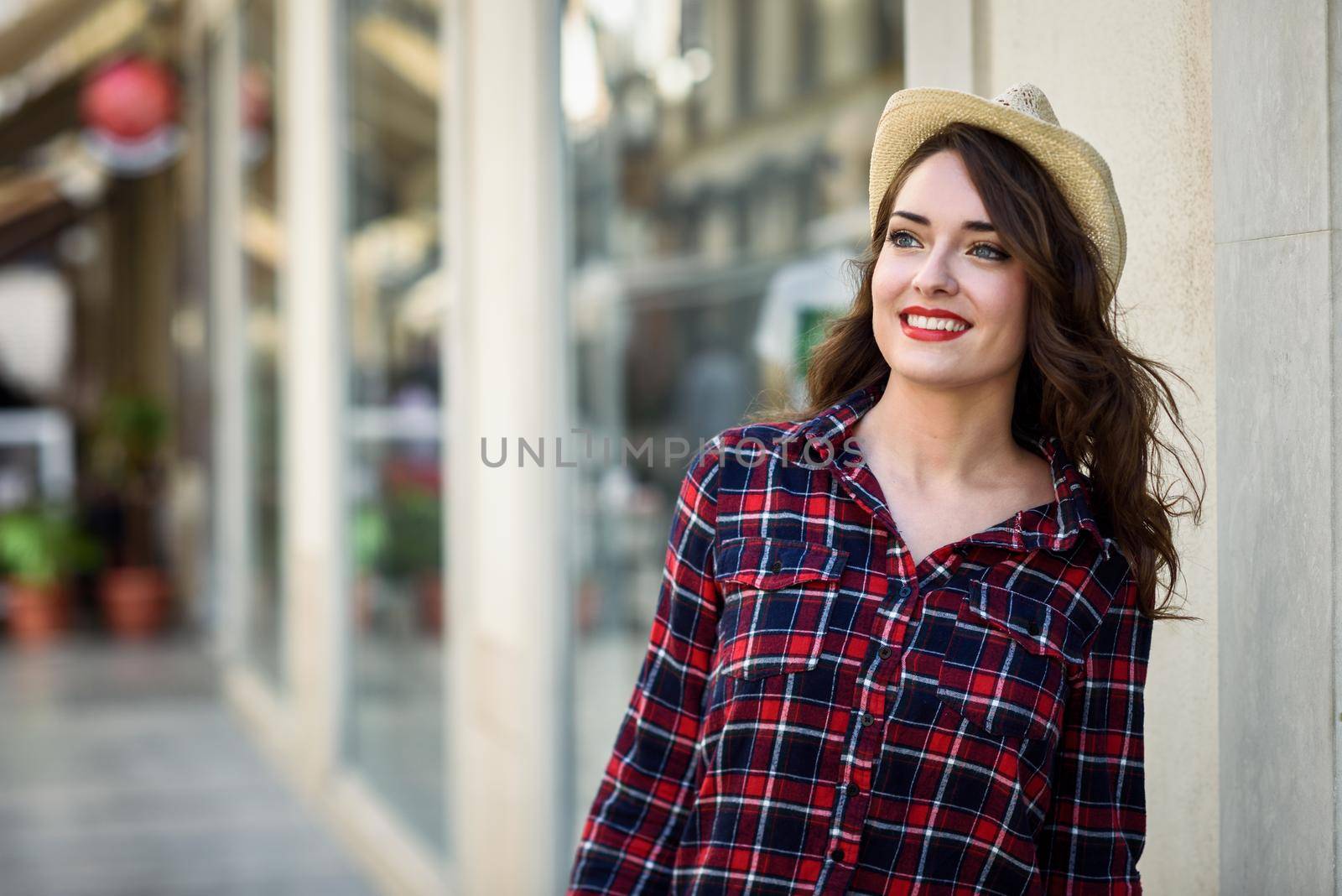 Young woman with beautiful blue eyes wearing plaid shirt and sun hat. Girl smiling in urban background.