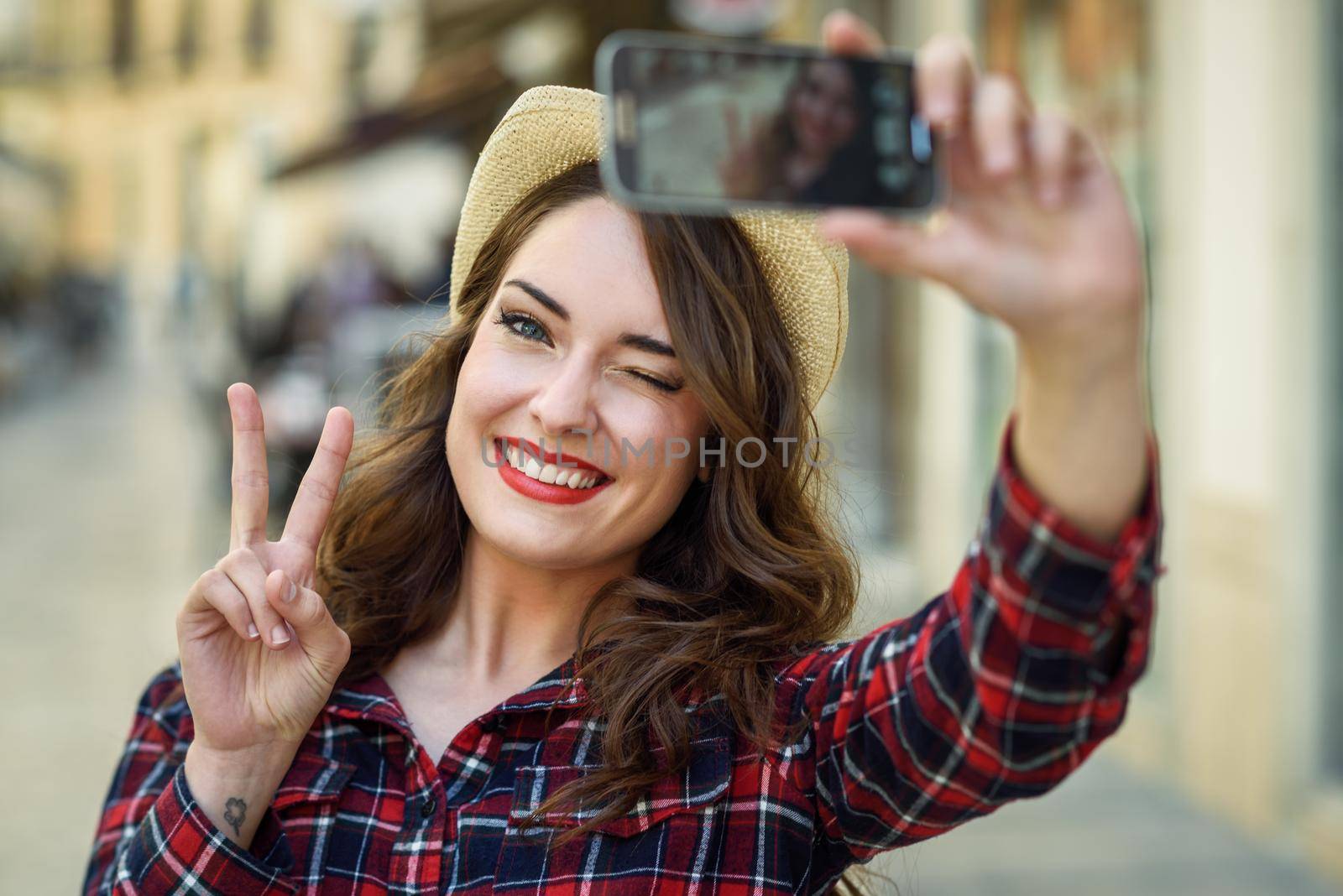 Portrait of a beautiful young woman selfie in the street with a smartphone. Young woman wearing plaid shirt and sun hat with red lips in urban background.