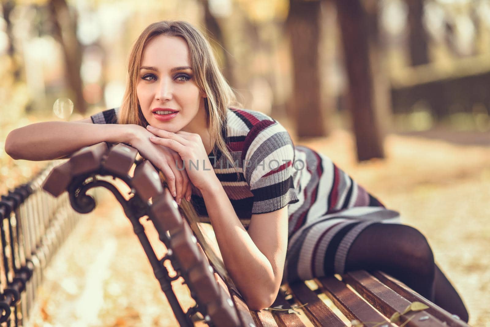 Pensive young blonde woman sitting on a bench in the street of a park with autumn colors. Beautiful girl in urban background wearing striped dress and black tights. Female with straight hair and blue eyes.
