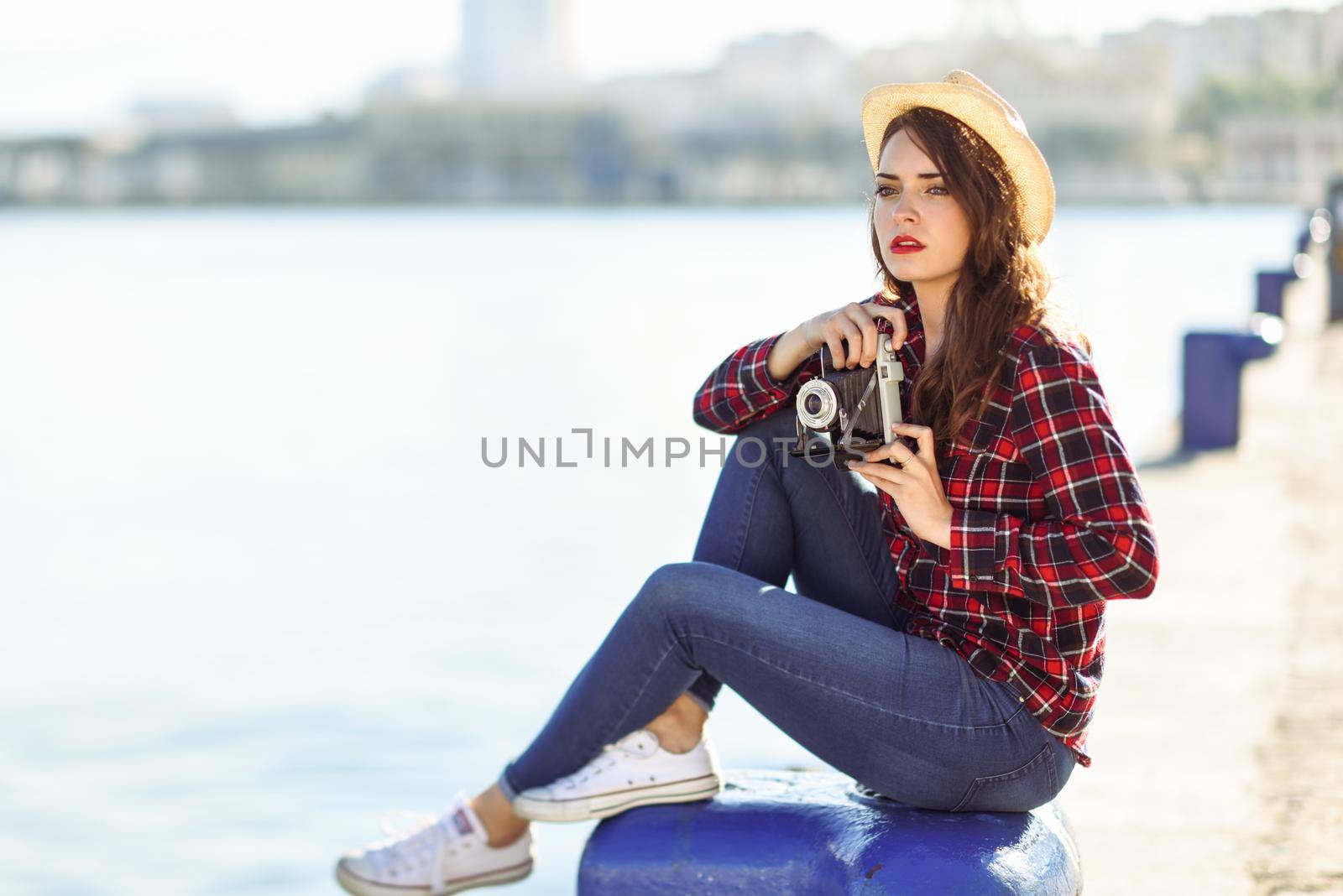 Young woman taking photographs with a vintage camera sitting in a harbour. Girl wearing plaid shirt, blue jeans and sun hat.