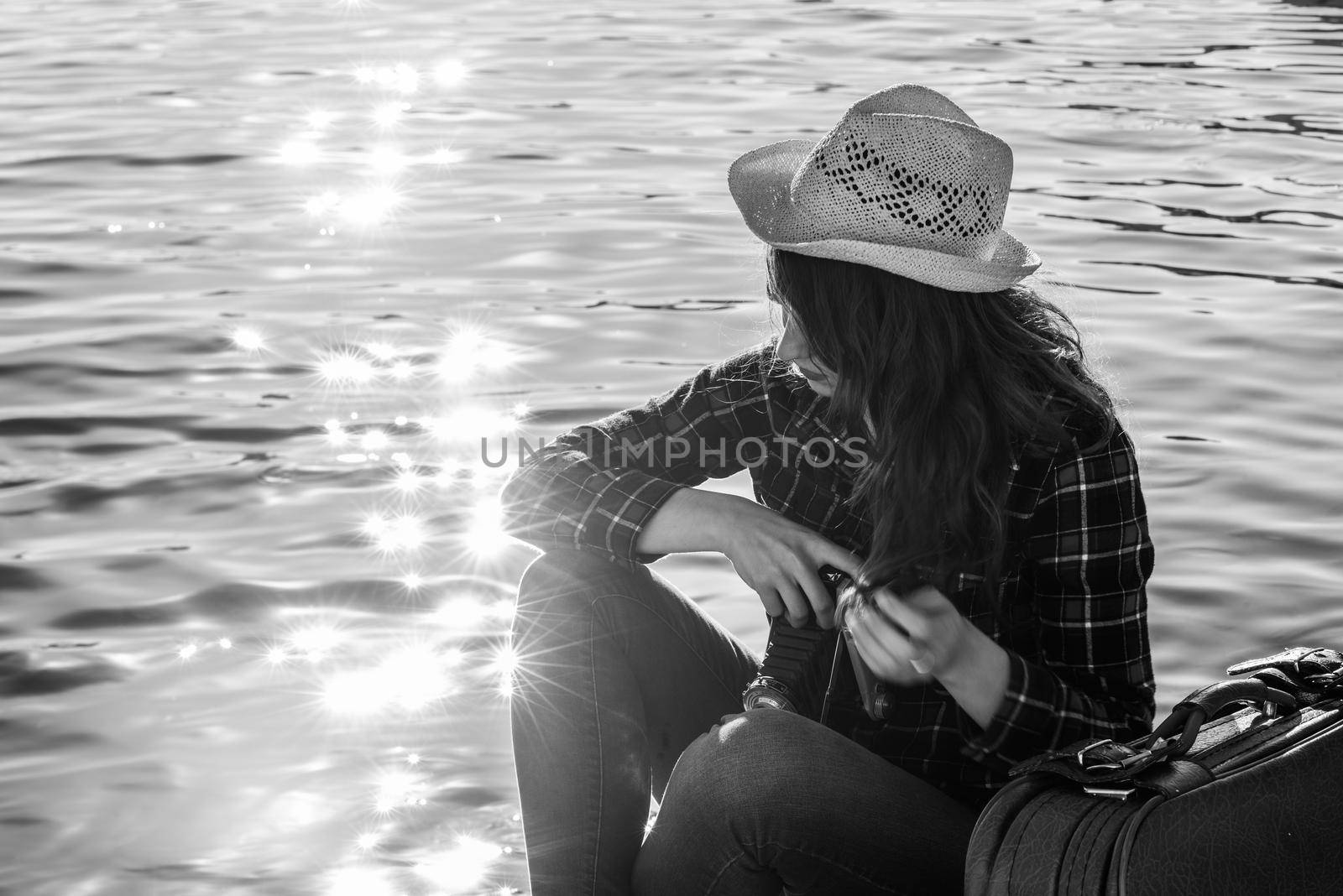Young woman taking photographs with a vintage camera sitting in a harbour. Girl wearing plaid shirt, blue jeans and sun hat. Black and white photograph.