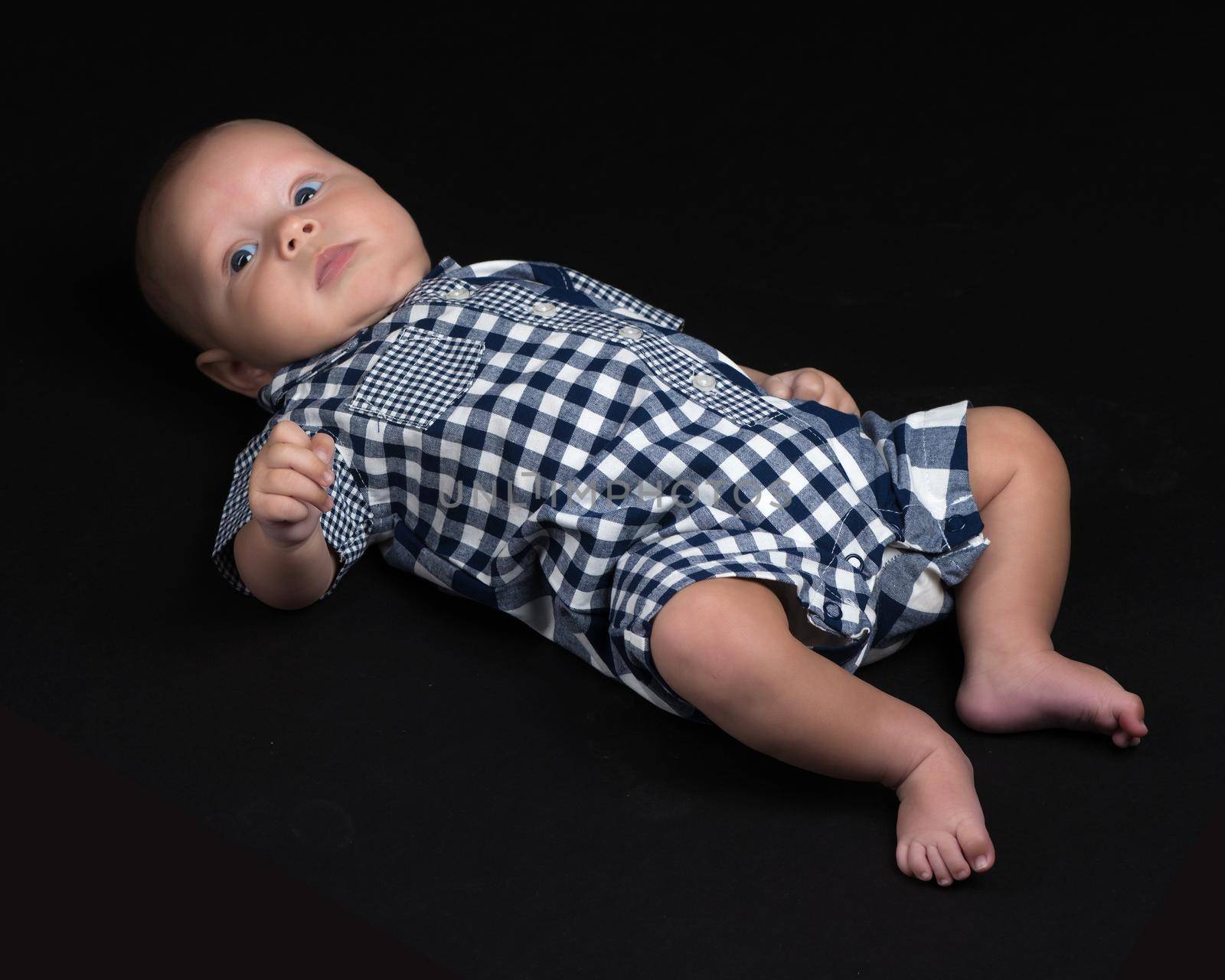 A charming baby lies on the blanket and looks into the camera on a black background. The concept of a happy childhood, the birth and upbringing of a child.