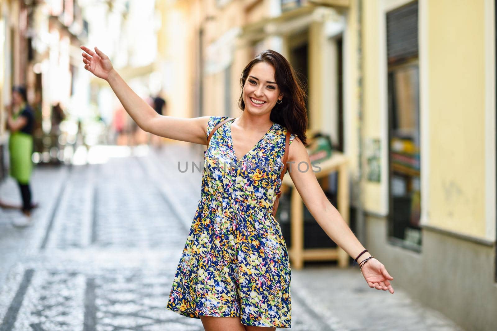 Happy young woman with blue eyes smiling outdoors. Girl wearing flower dress raising arms in urban background. Happiness concept.