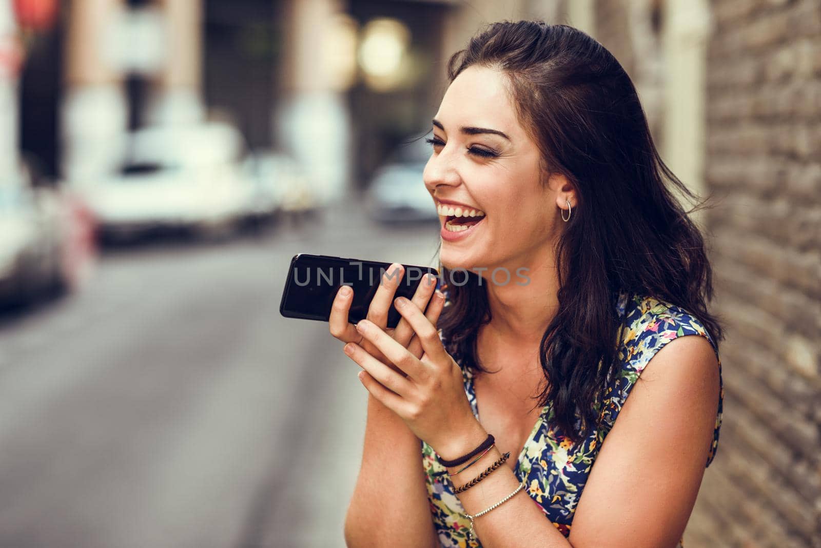 Smiling young woman recording voice note in her smart phone outdoors. Girl wearing flower dress in urban background. Technology concept.