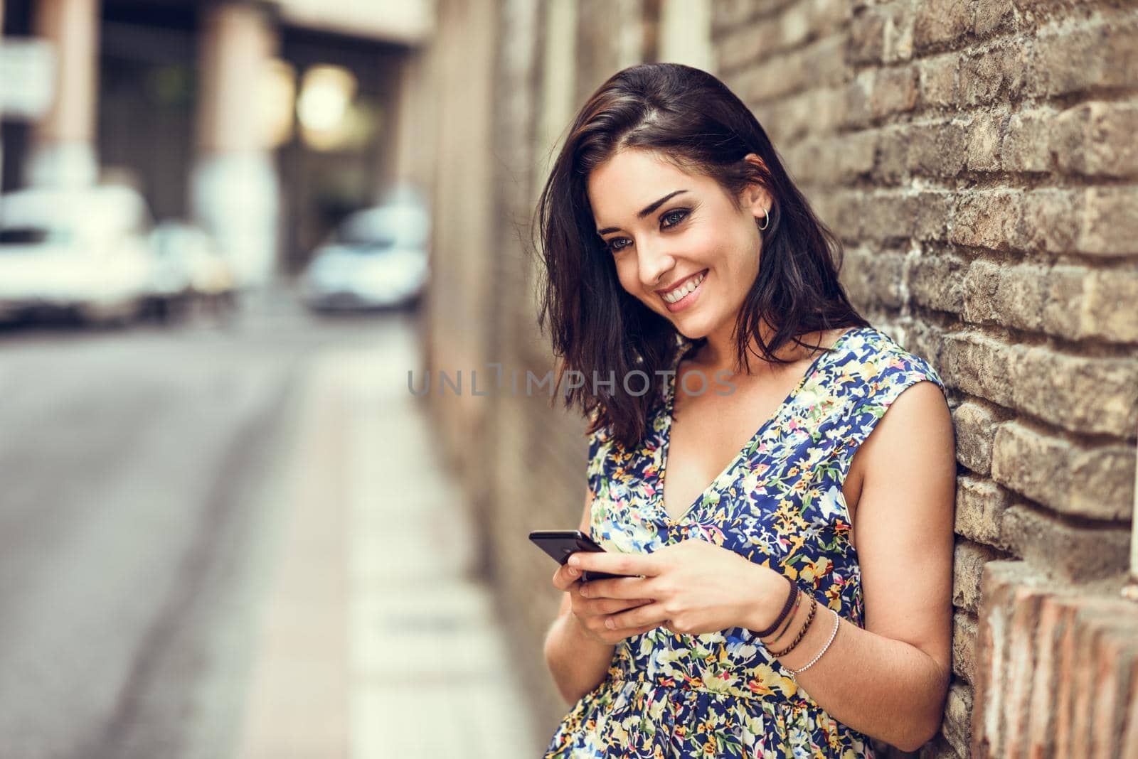 Smiling young woman using her smart phone outdoors. Girl wearing flower dress in urban background. Technology concept.