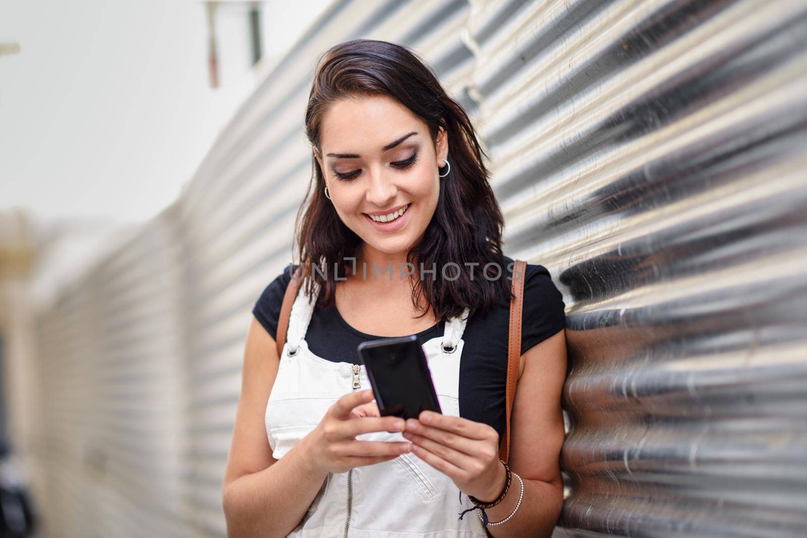 Smiling young woman texting with her smart phone outdoors. Girl wearing denim dress in urban background. Technology concept.