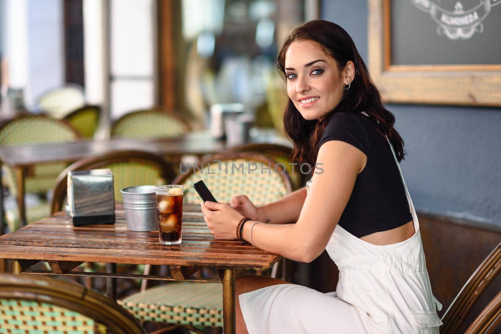 Beautiful girl with blue eyes sitting on urban cafe using smart phone. Young woman with brown wavy hairstyle wearing white denim dress. Lifestyle concept.
