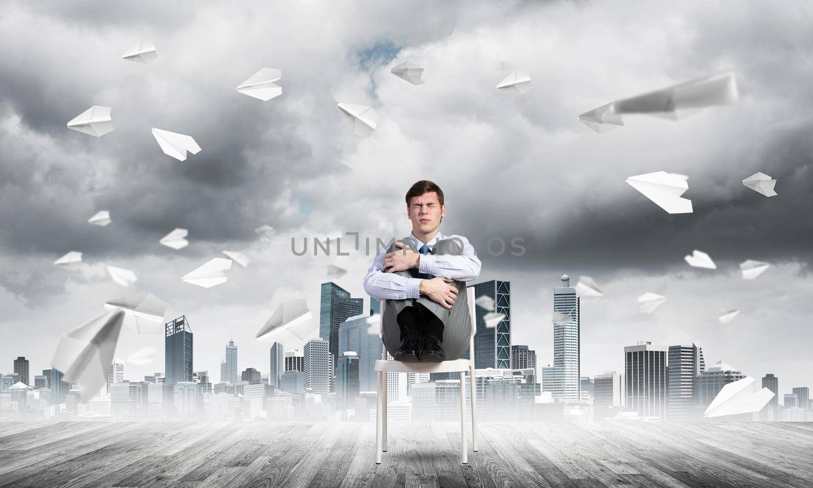 scared businessman sitting on a chair with his hands clasped his knees, paper plane fly around