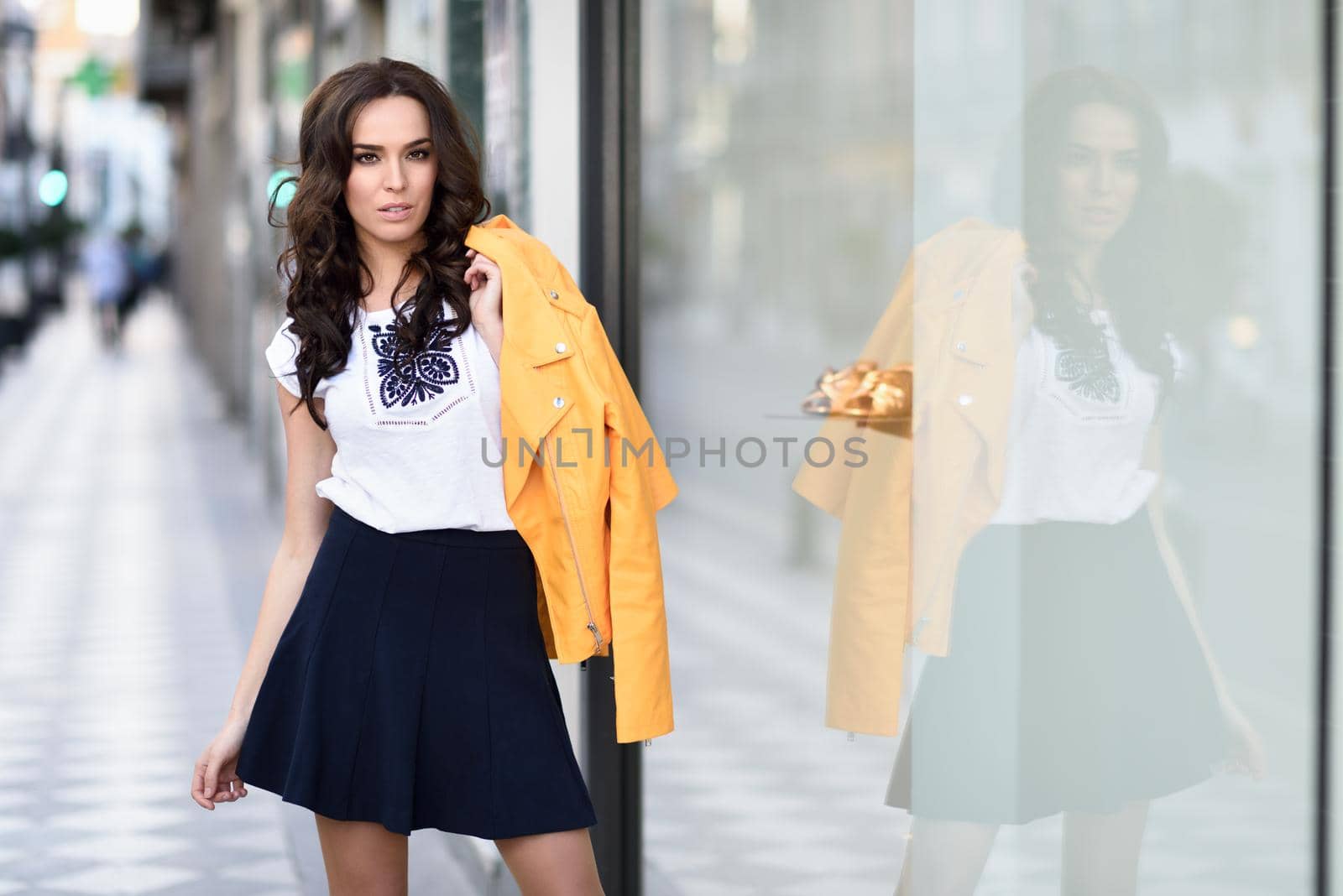 Young brunette woman, model of fashion, wearing orange modern jacket and blue skirt, standing in urban background.