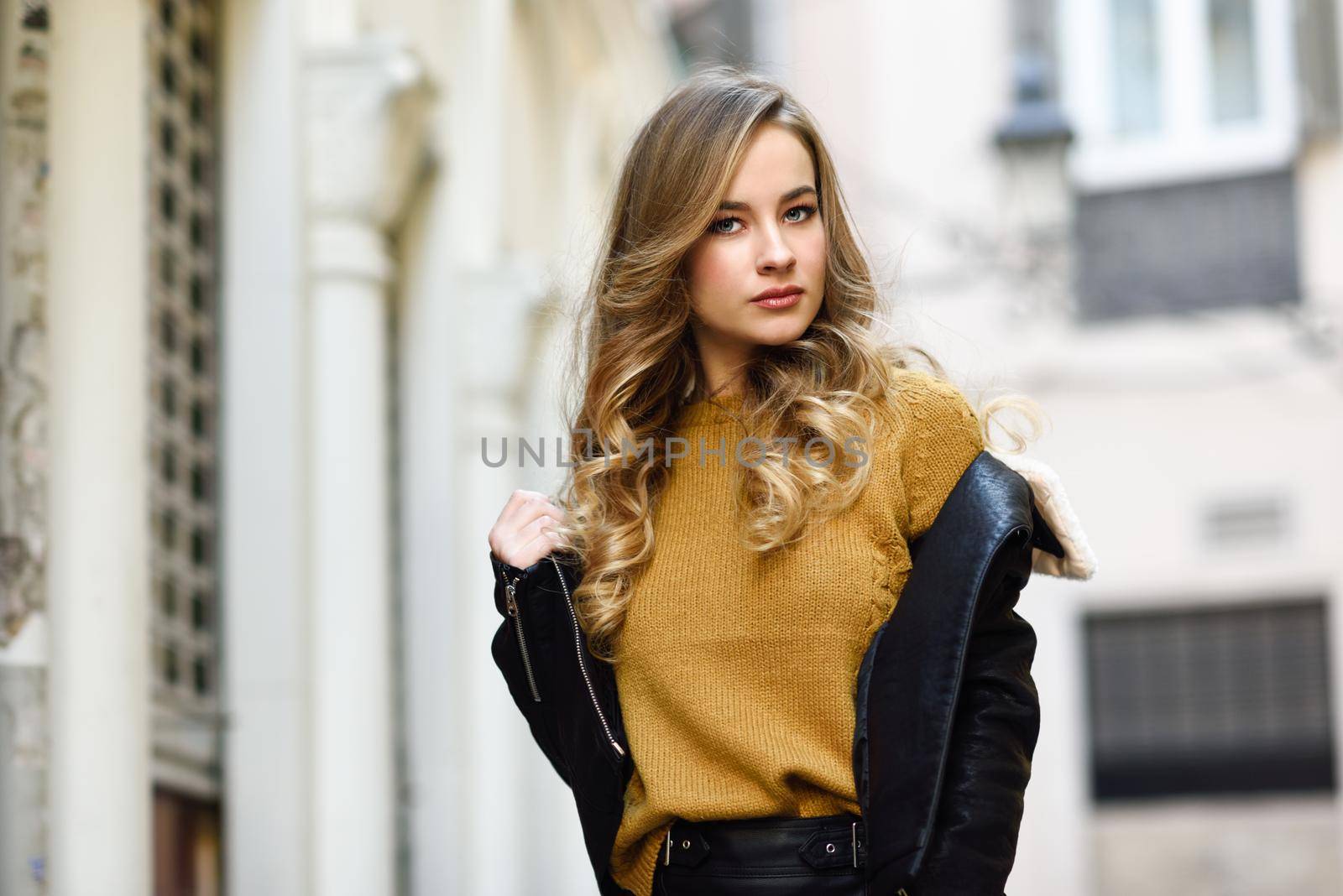Blonde woman in urban background. Beautiful young girl wearing black leather jacket and mini skirt standing in the street. Pretty russian female with long wavy hair hairstyle and blue eyes.