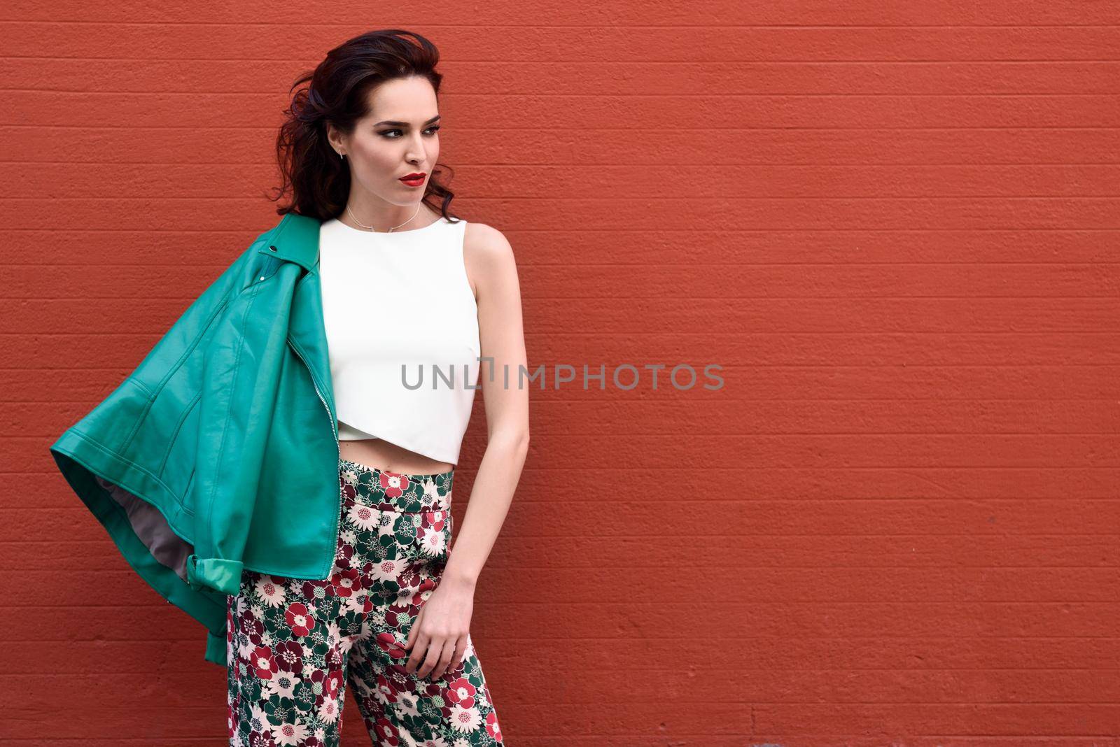 Young brunette woman, model of fashion, wearing green modern jacket and flower pants on red wall. Pretty caucasian girl with long wavy hairstyle. Female with red lips in urban background.