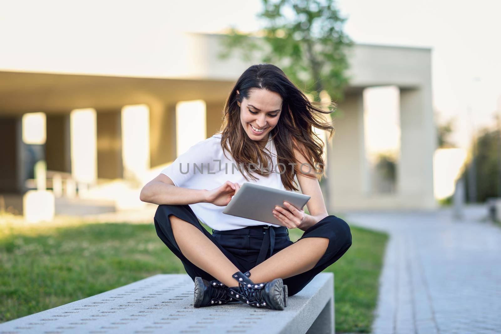 Young woman using digital tablet sitting outdoors in urban background. by javiindy