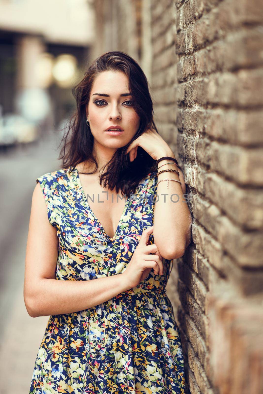 Girl with blue eyes standing next to brick wall outdoors. Young woman in her twenties wearing flower dress in urban background. Beauty and fashion concept.