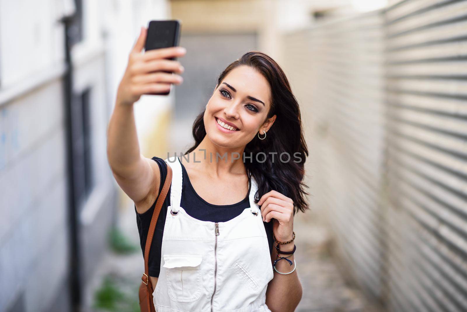 Young woman taking selfie photograph with her smart phone outdoors. Girl wearing denim dress in urban background. Technology concept.