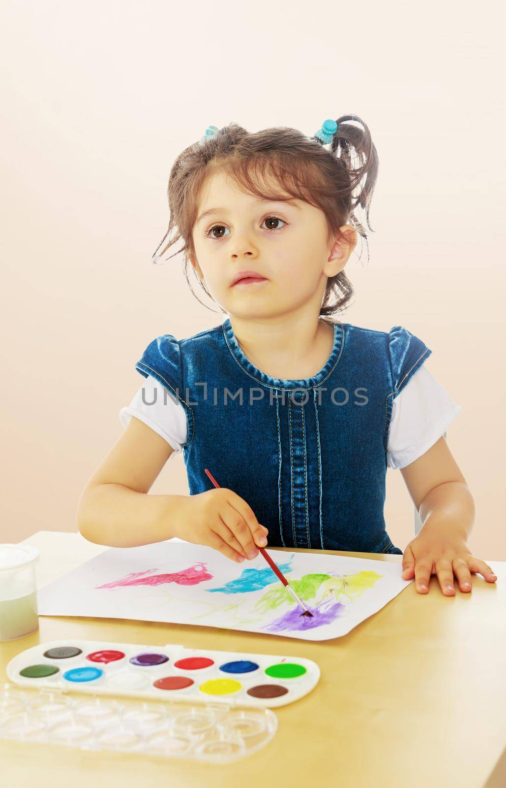 Cute little girl draws with watercolors on a sheet of white paper . Girl sitting at the table.On a brown gradient background.