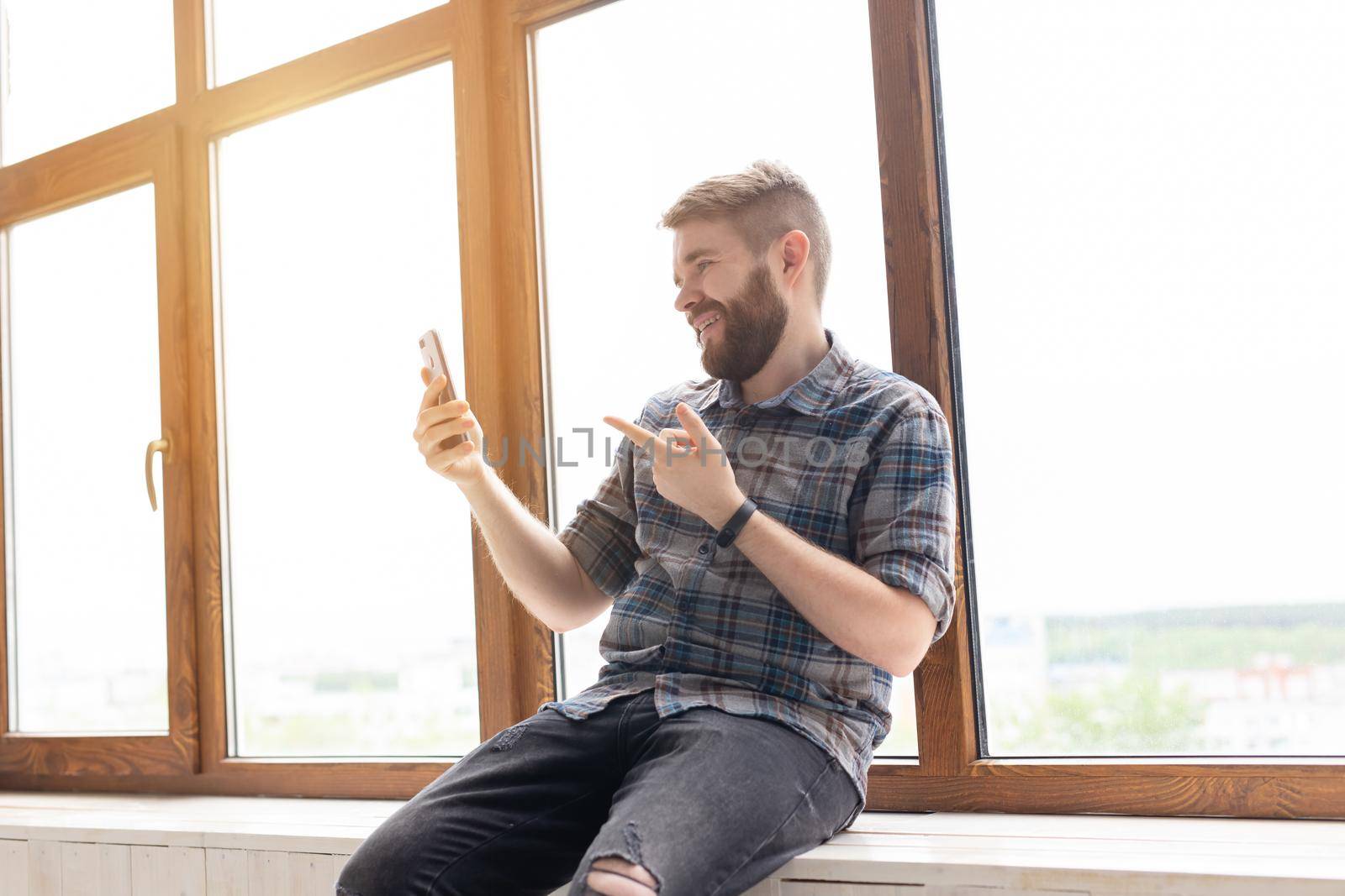 Cute young positive guy hipster with a beard is sitting on the windowsill by the big window and talking on the video communication using a smartphone and wireless high-speed Internet. by Satura86