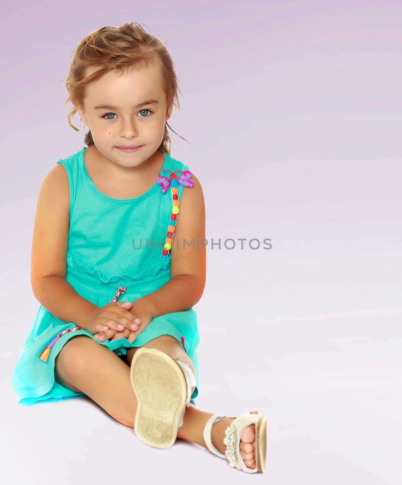 Beautiful little blond girl dressed in a blue dress. Girl sitting on the floor putting one leg over the other.Not a purple gradient background.
